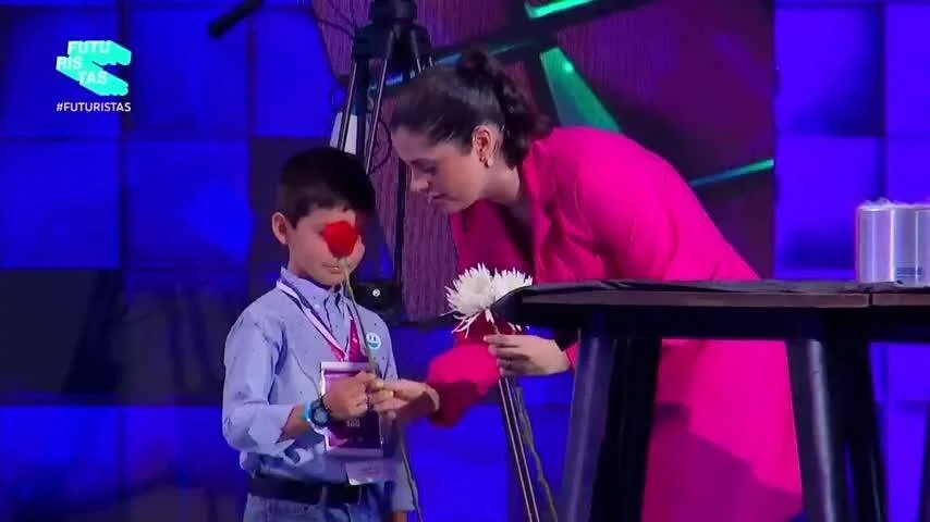 Una mujer se inclina hacia un niño con nariz de payaso y credencial durante una presentación en escenario del programa #Futuristas.