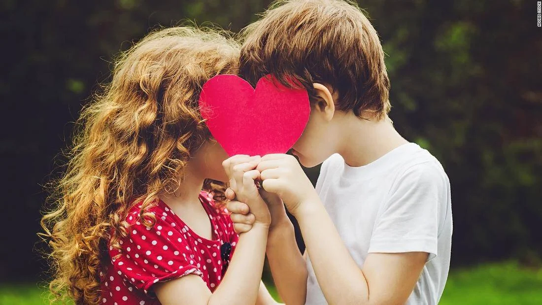 Dos niños se acercan ocultando sus rostros detrás de un corazón de papel rojo que sostienen juntos.