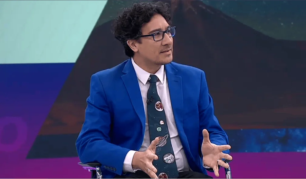 Hombre con blazer azul, corbata con planetas y gafas habla gesticulando en un estudio de televisión.