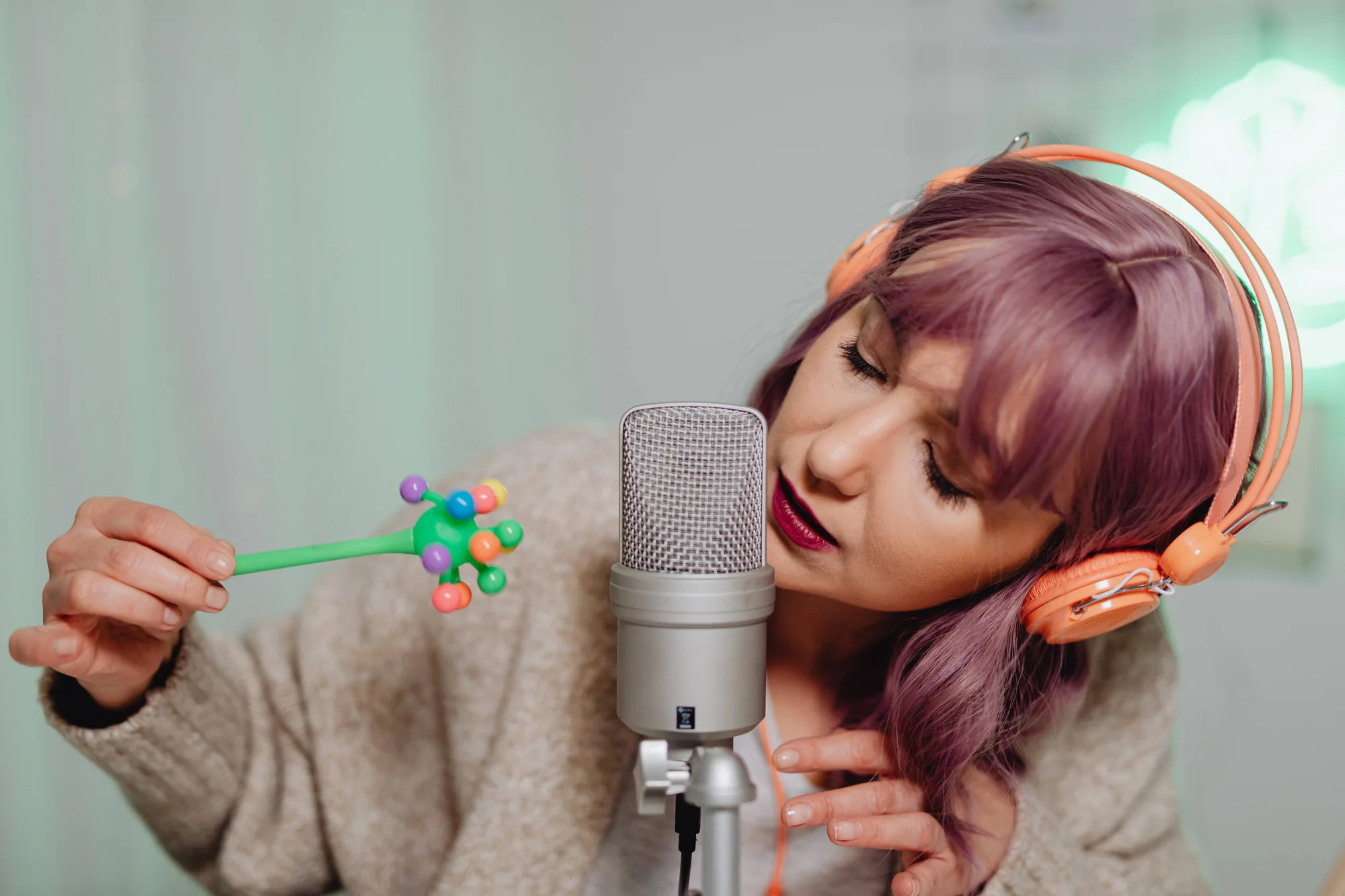 Mujer de cabello morado con auriculares naranjas acerca un sonajero colorido a un micrófono de condensador durante una sesión de grabación ASMR.
