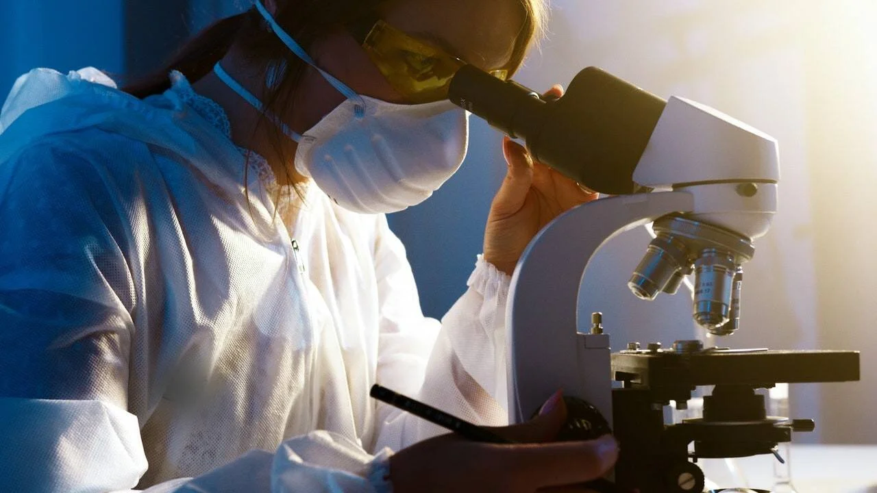 Investigadora con bata, mascarilla y gafas de protección observa muestras a través de un microscopio en un laboratorio.