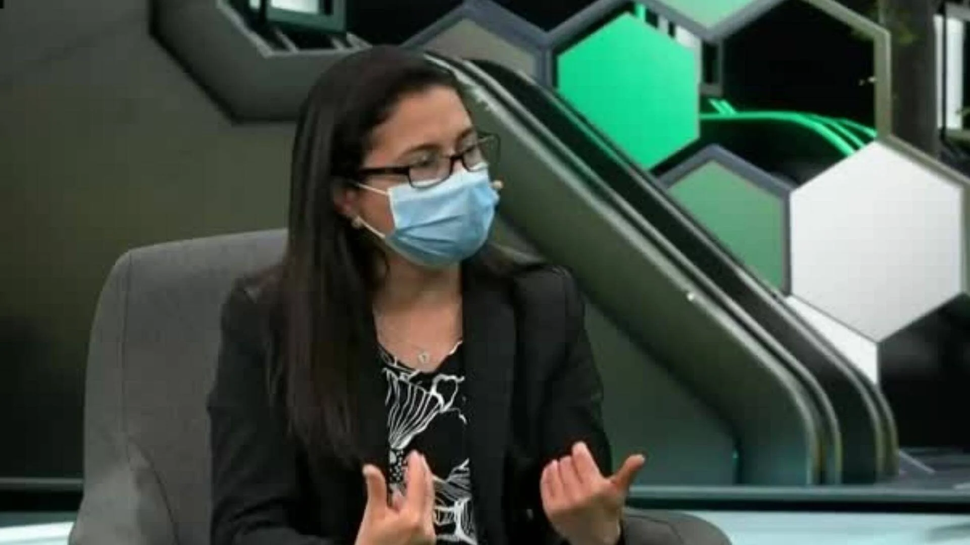 Mujer con anteojos y barbijo quirúrgico habla gesticulando con las manos en un estudio de televisión con decorado hexagonal verde.