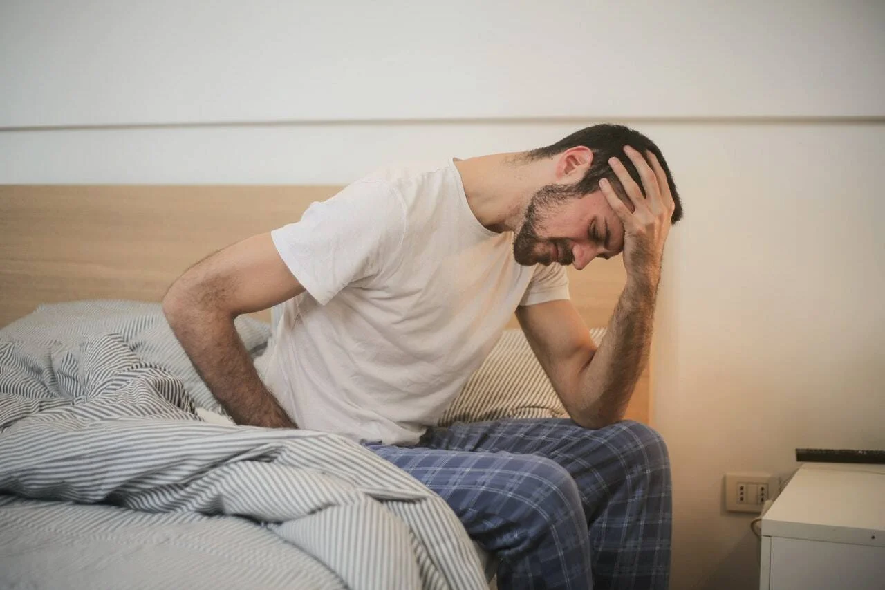Hombre en pijama sentado al borde de la cama sosteniéndose la cabeza con gesto de dolor o agotamiento.