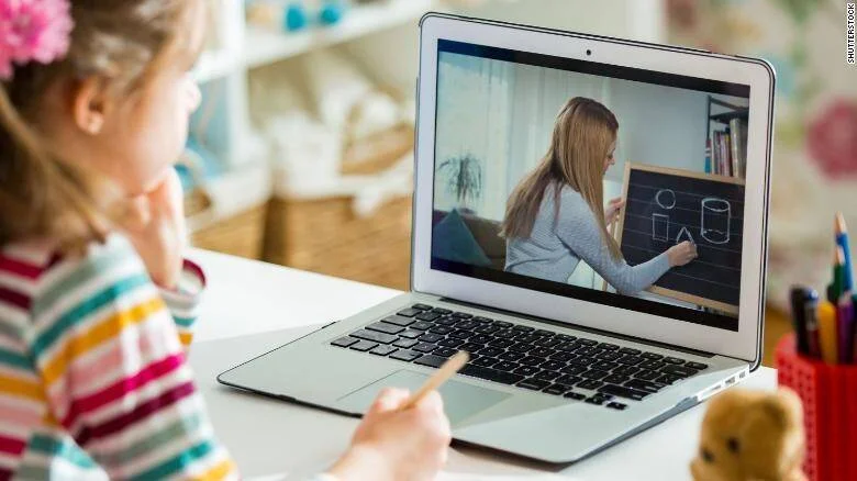 Una niña pequeña observa a su maestra enseñar figuras geométricas en una pizarra a través de una videollamada en su laptop.
