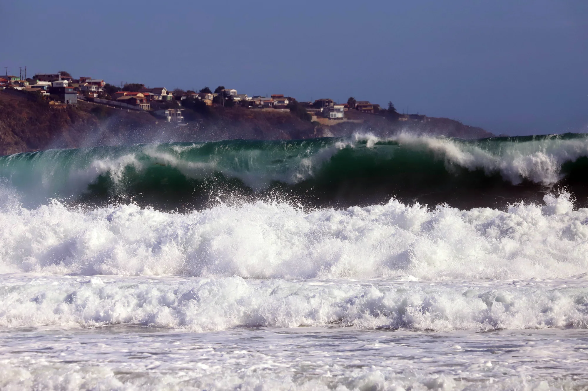 Grandes olas verdes rompen con fuerza cerca de la orilla, con casas sobre un acantilado al fondo.