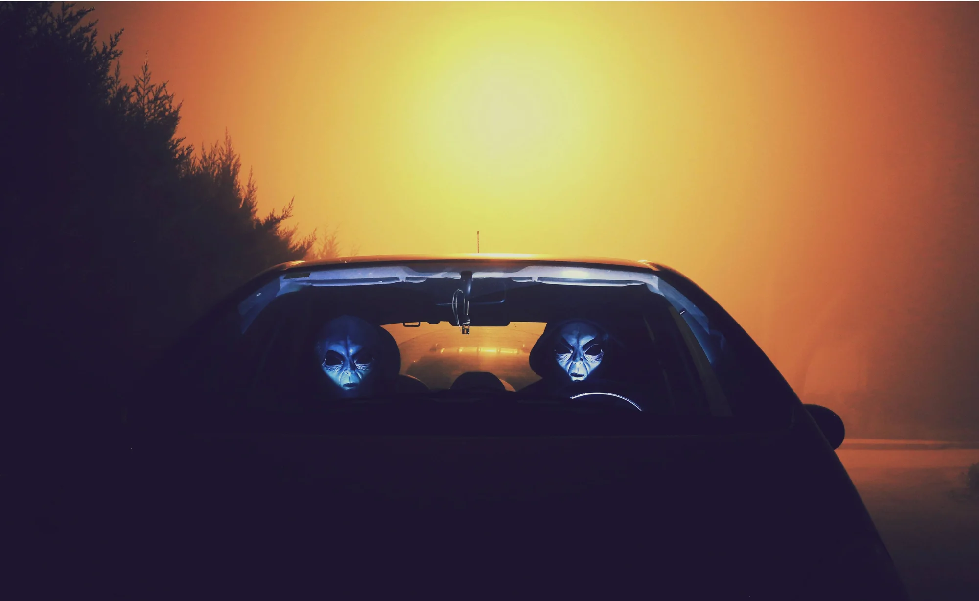 Dos personas con máscaras de extraterrestres sentadas en un auto visto desde atrás, con un resplandor anaranjado al fondo.