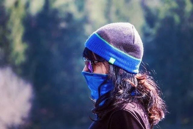 Mujer de perfil con gorro de lana azul, gafas y cubrebocas azul en entorno boscoso.