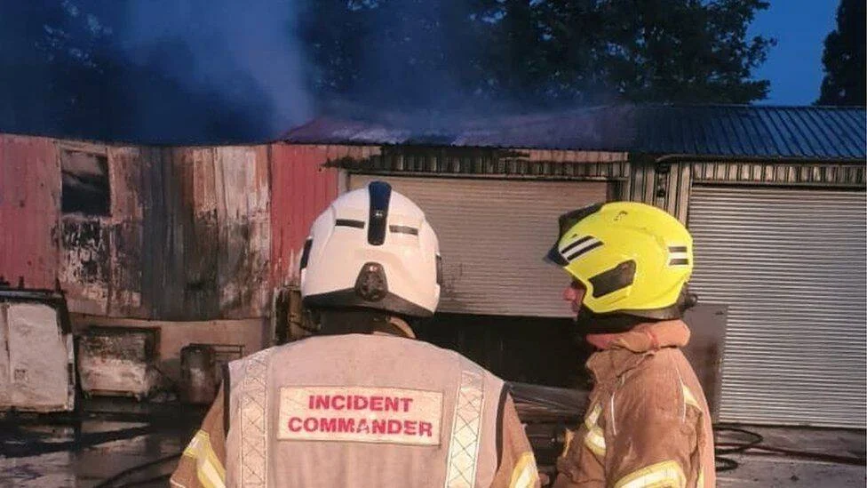 Dos bomberos, uno con chaleco de comandante de incidentes, observan un almacén industrial envuelto en humo y fuego.