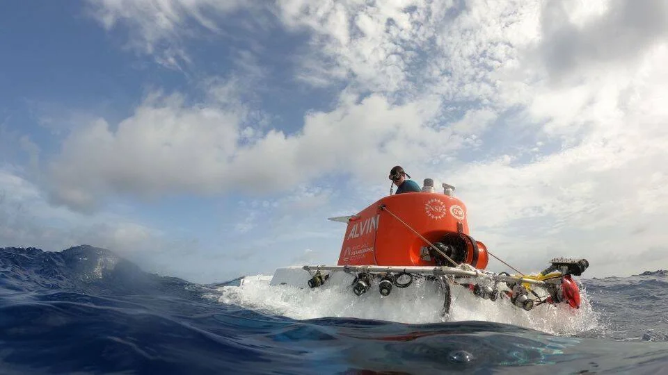 El submarino de investigación Alvin, con cúpula naranja, emerge en la superficie del océano con una persona sobre él.