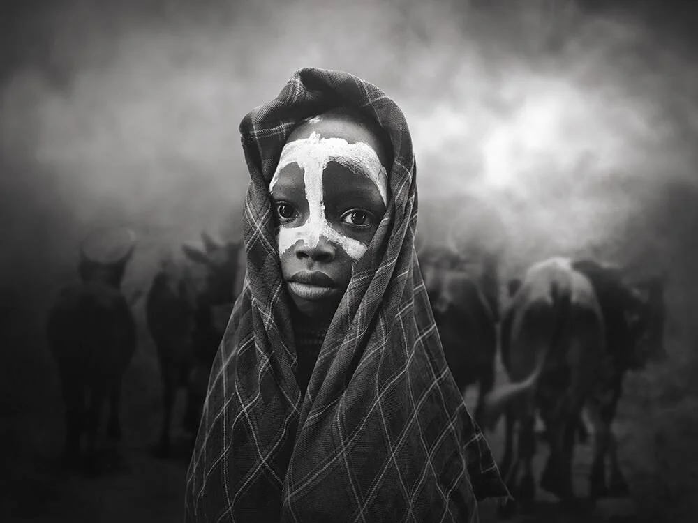 Niño con pintura blanca ritual en el rostro y manta a cuadros, rodeado de ganado entre el humo, en blanco y negro.