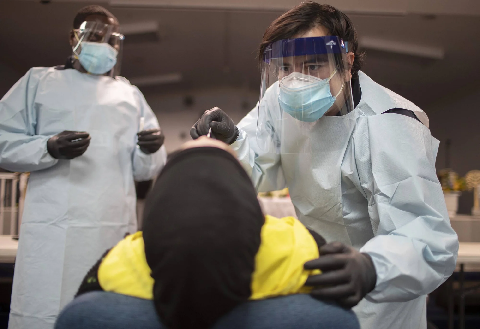 Profesional de salud con EPP completo toma una muestra nasal a un paciente mientras otro trabajador observa al fondo.