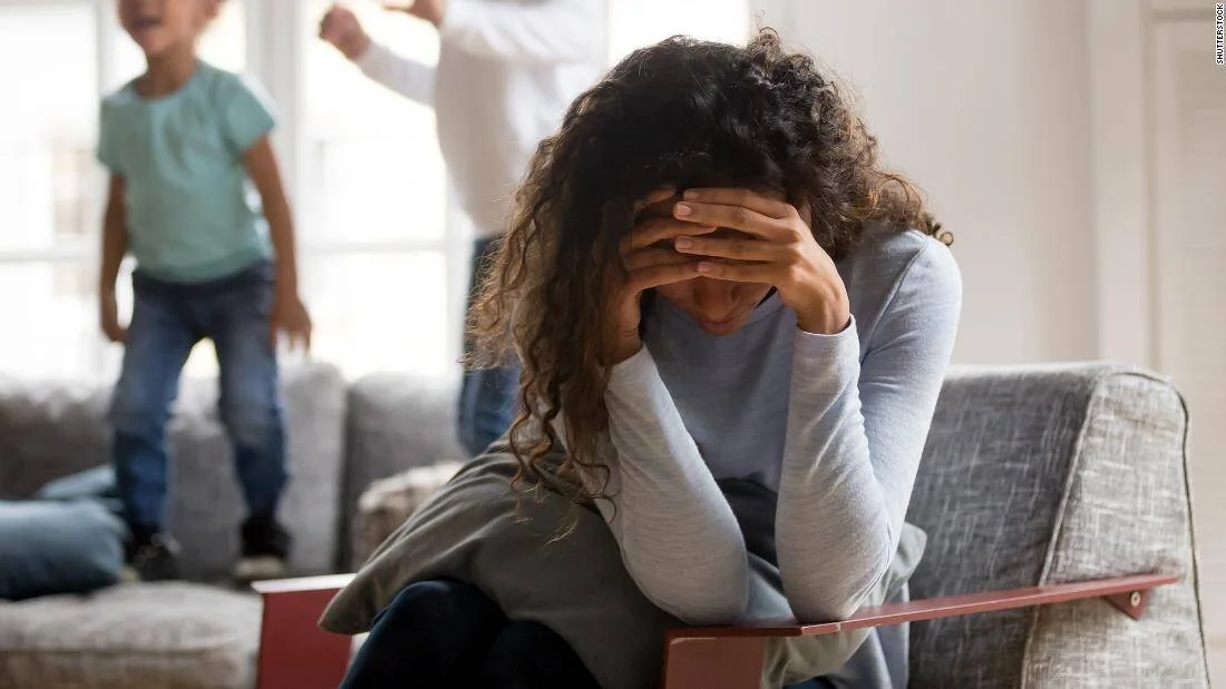 Mujer con la cabeza entre las manos, sentada en un sillón, mientras un niño salta en el sofá al fondo.