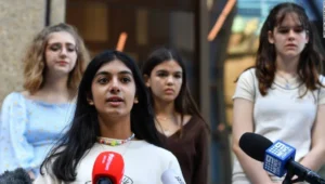 Una joven habla frente a micrófonos de canales de noticias australianos, acompañada por tres adolescentes detrás de ella.