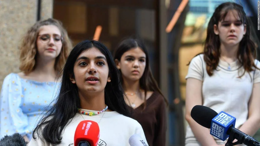 Una joven habla frente a micrófonos de canales de noticias australianos, acompañada por tres adolescentes detrás de ella.