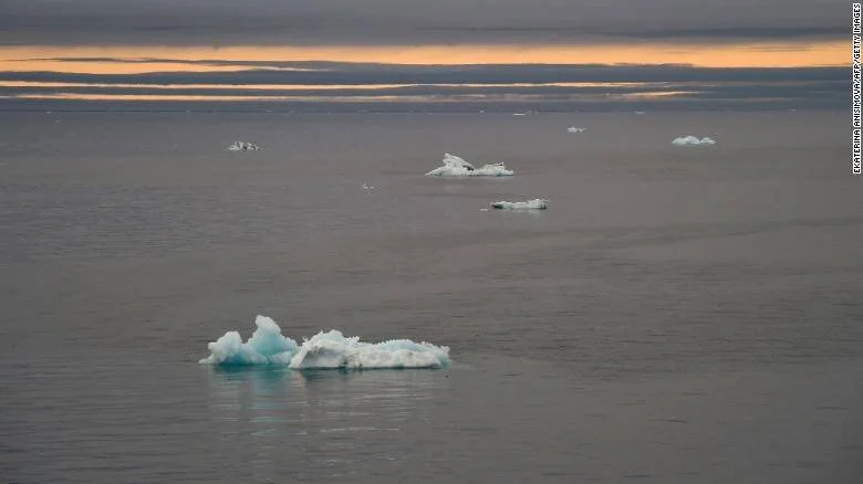 Pequeños bloques de hielo a la deriva en un mar ártico gris bajo un cielo de atardecer anaranjado, evidenciando el deshielo polar.