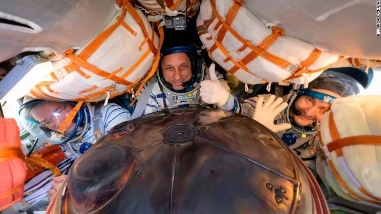 Tres astronautas con trajes espaciales sonríen dentro de la cápsula Soyuz tras su regreso a Tierra; el del centro hace señal de pulgar arriba.