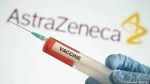 Mano con guante azul sostiene una jeringa etiquetada 'Vaccine' con el logo de AstraZeneca al fondo.