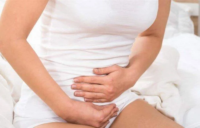Mujer vestida de blanco sostiene su abdomen con ambas manos, indicando dolor o malestar estomacal.