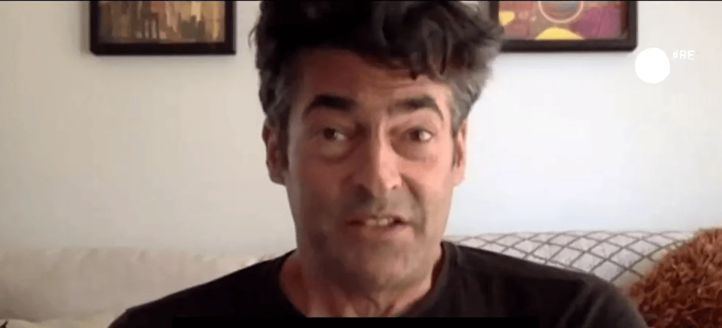 Hombre de mediana edad con cabello oscuro canoso habla frente a cámara en un interior con cuadros en la pared.
