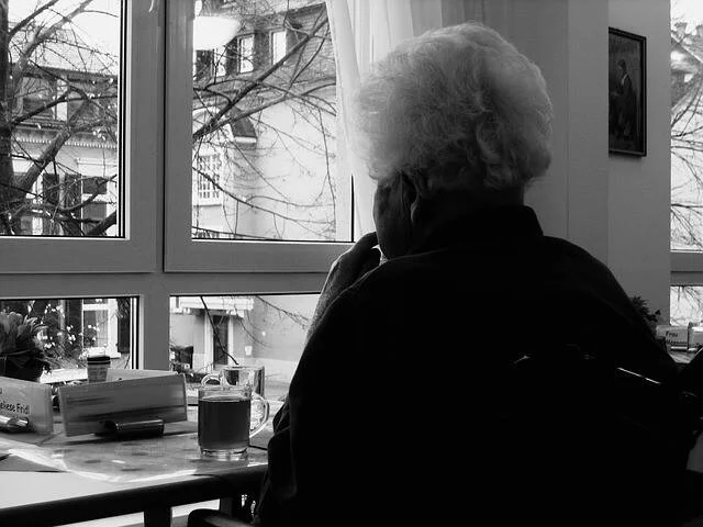 Mujer mayor de espaldas contempla la calle a través de una ventana, sentada ante una mesa con una taza.
