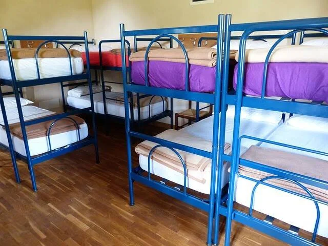 Habitación de albergue con varias literas de metal azul tendidas con sábanas de colores sobre suelo de parqué.