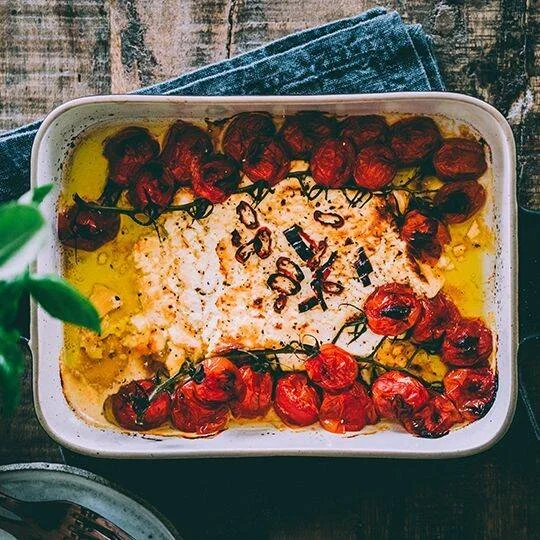Bloque de queso feta horneado con tomates cherry asados y aceite de oliva en una fuente rectangular.