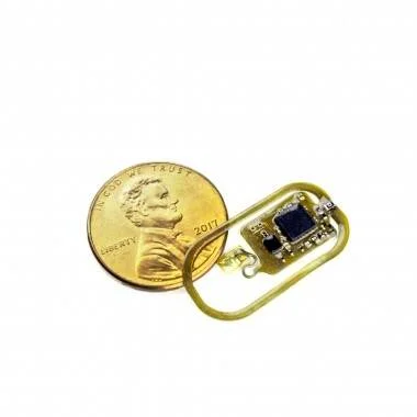 Dispositivo electrónico miniaturizado con componentes integrados, colocado junto a una moneda de un centavo estadounidense para mostrar su tamaño reducido.