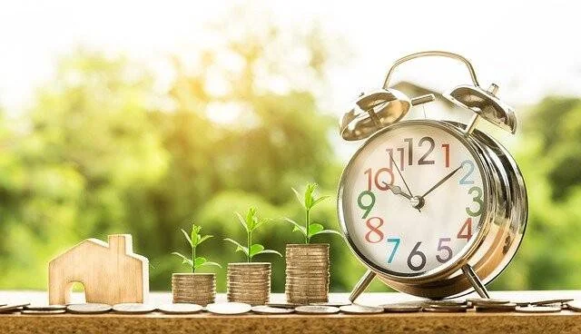Pilas de monedas en aumento con plantas brotando, una casa de madera y un reloj despertador, simbolizando inversión y crecimiento financiero.