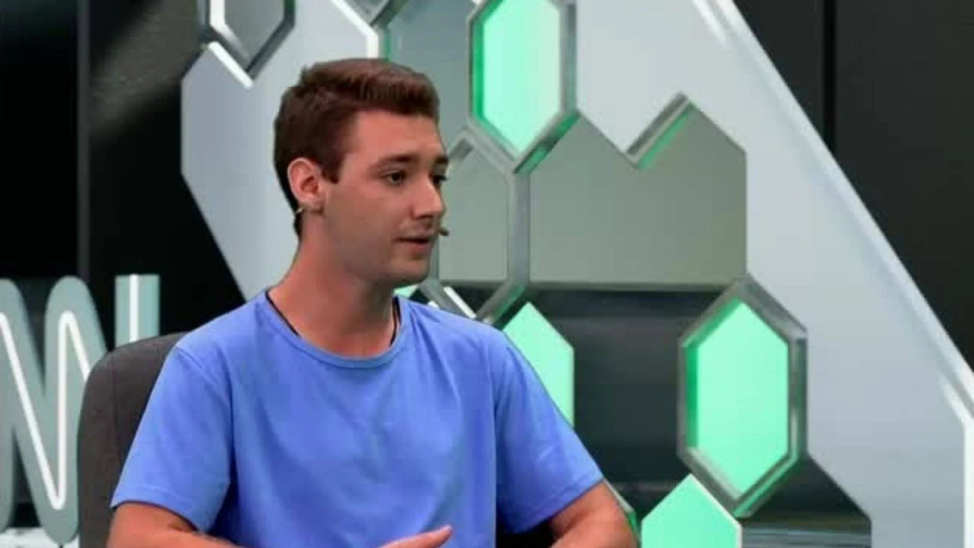 Hombre joven con remera azul habla en un estudio de televisión con decorado de hexágonos iluminados en verde.