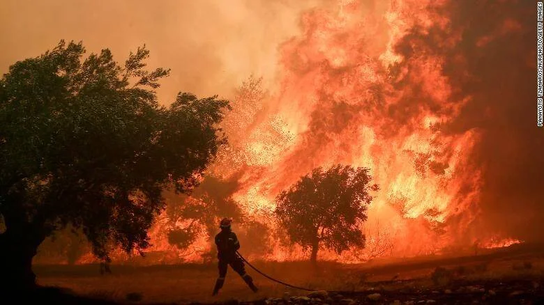 Un bombero sostiene una manguera frente a una enorme pared de fuego que arrasa árboles durante un incendio forestal.