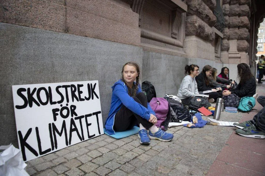 Joven sentada en la acera junto a un cartel que dice 'Skolstrejk för Klimatet' (Huelga escolar por el clima), acompañada de otros estudiantes.