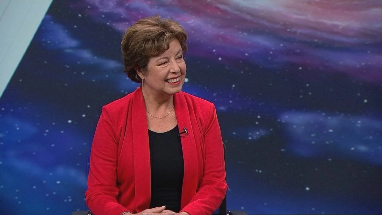 Mujer de mediana edad con saco rojo sonríe durante una entrevista en un estudio de televisión con fondo de galaxia.
