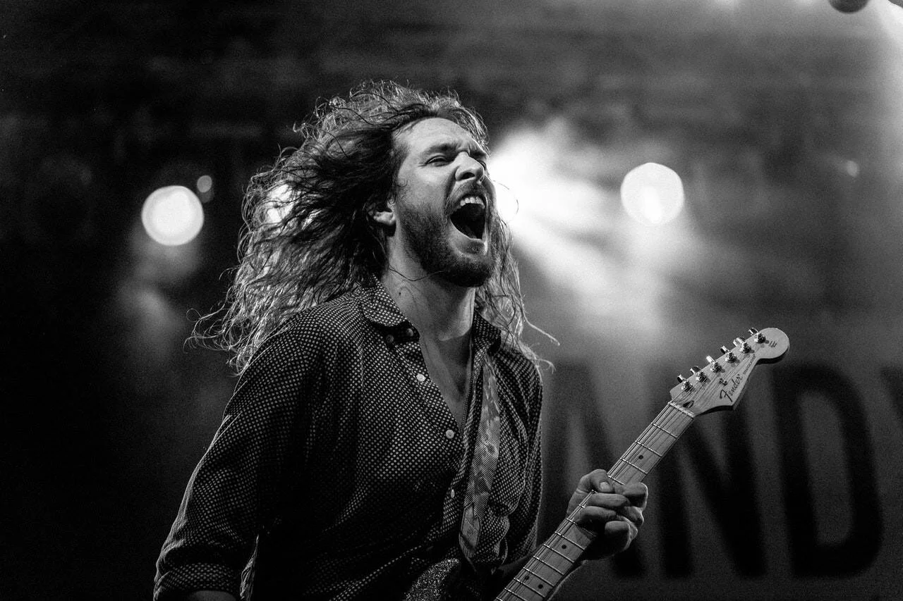 Guitarrista de cabello largo grita con intensidad mientras toca una Fender en un concierto iluminado por focos.