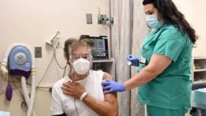 Una enfermera con guantes aplica una inyección en el brazo a un paciente mayor con mascarilla sentado en una camilla médica.