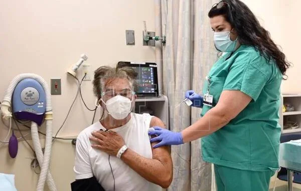 Una enfermera con guantes aplica una inyección en el brazo a un paciente mayor con mascarilla sentado en una camilla médica.