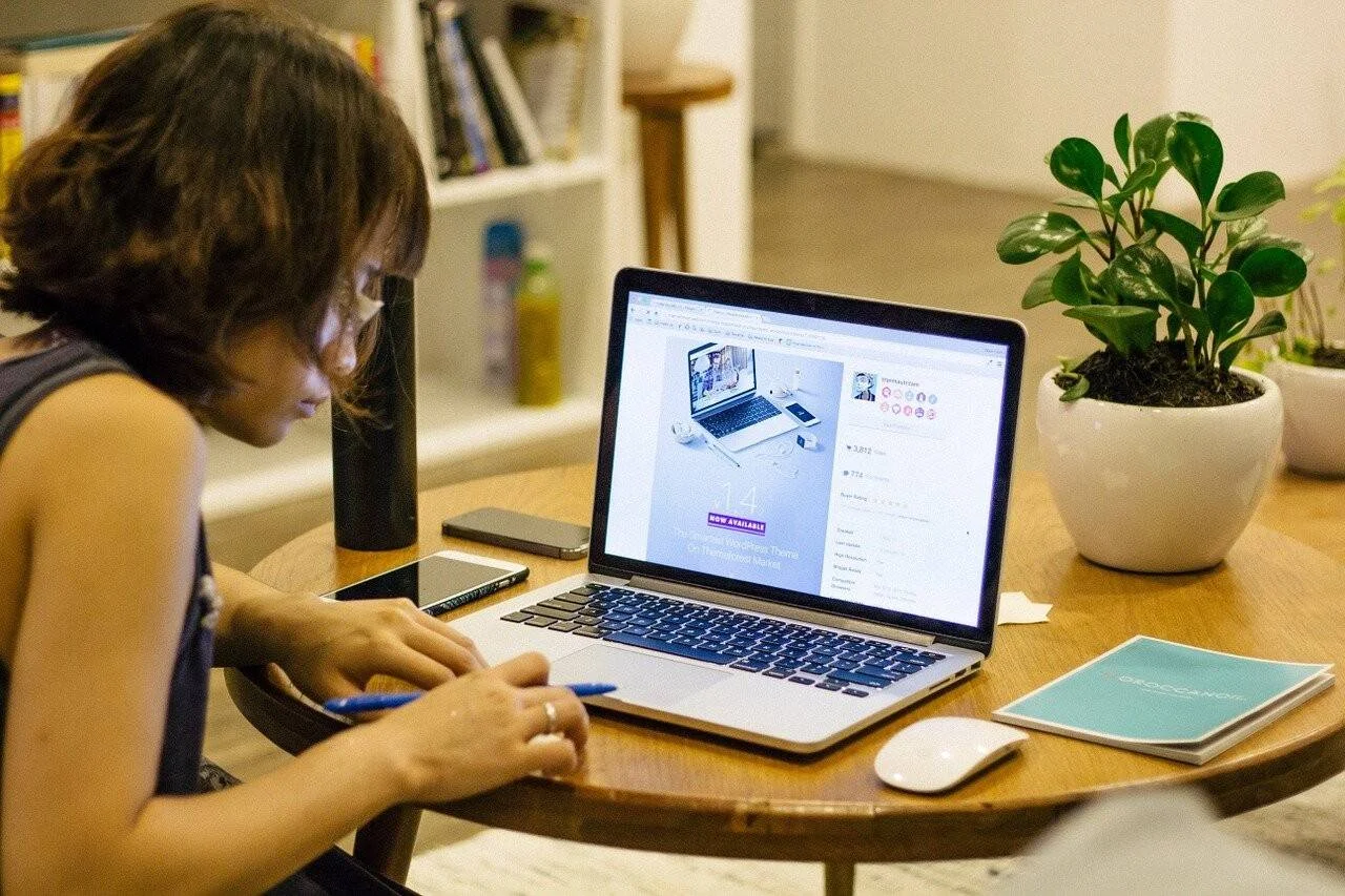 Mujer con gafas toma notas frente a una laptop abierta en una mesa redonda de madera con mouse y planta decorativa.