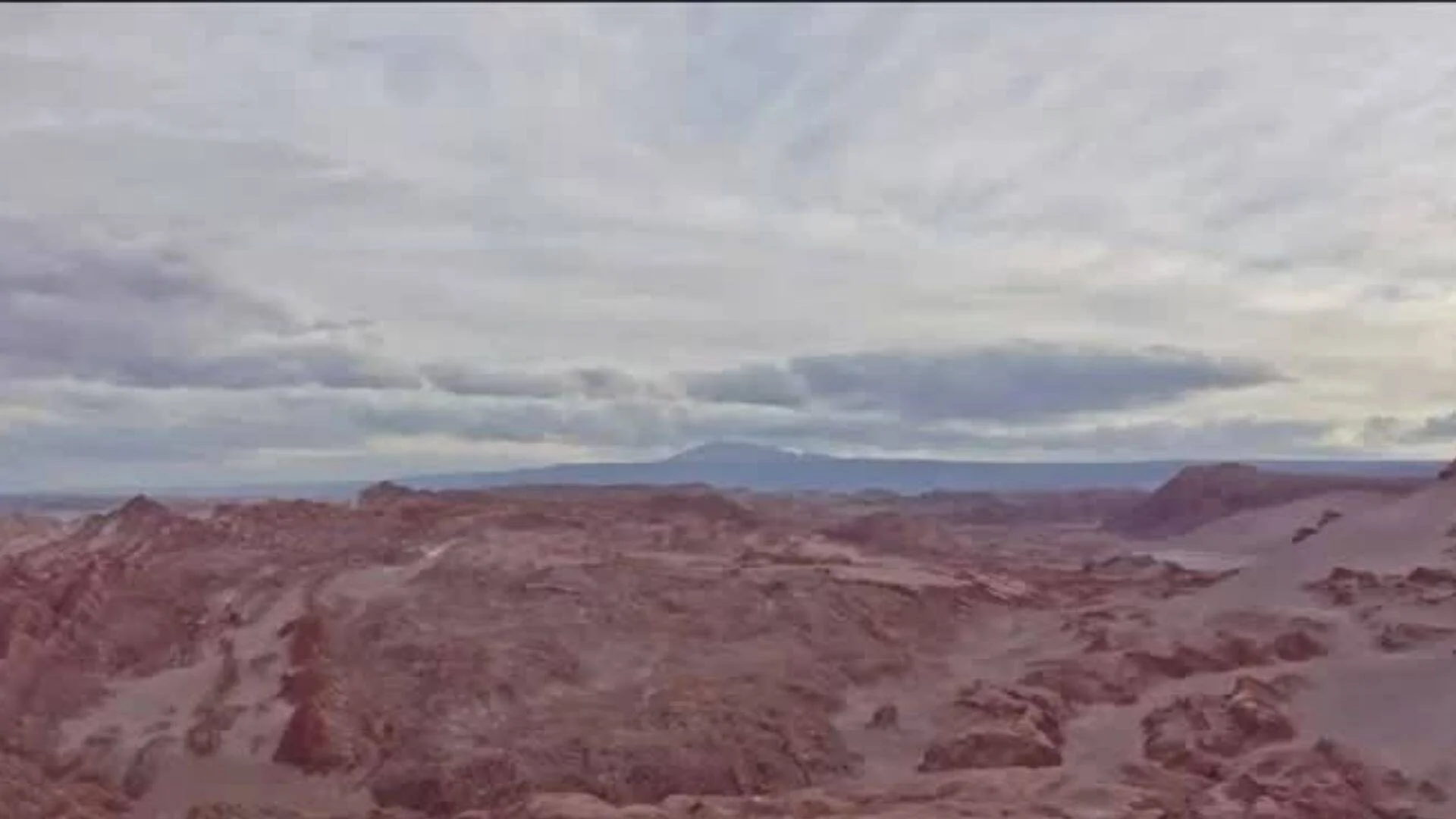 Vasta llanura desértica de suelo rojizo y rocoso bajo un cielo nublado, con montañas en el horizonte.