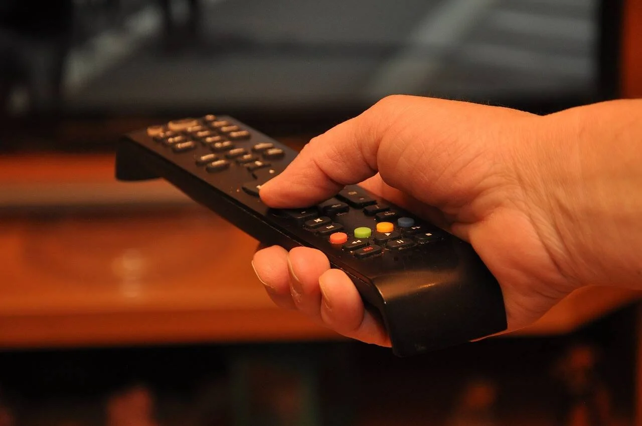 Una mano sostiene un control remoto negro apuntando hacia un televisor en segundo plano.