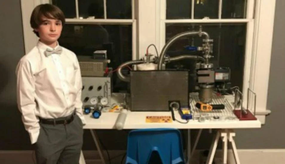 Un niño con camisa blanca y corbatín posa junto a un reactor de fusión nuclear construido en casa, con equipos científicos sobre una mesa.