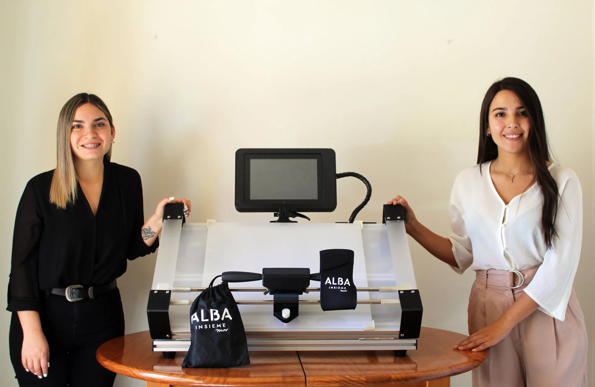 Dos mujeres sonríen junto a un dispositivo tecnológico de la marca Alba Insieme sobre una mesa de madera.