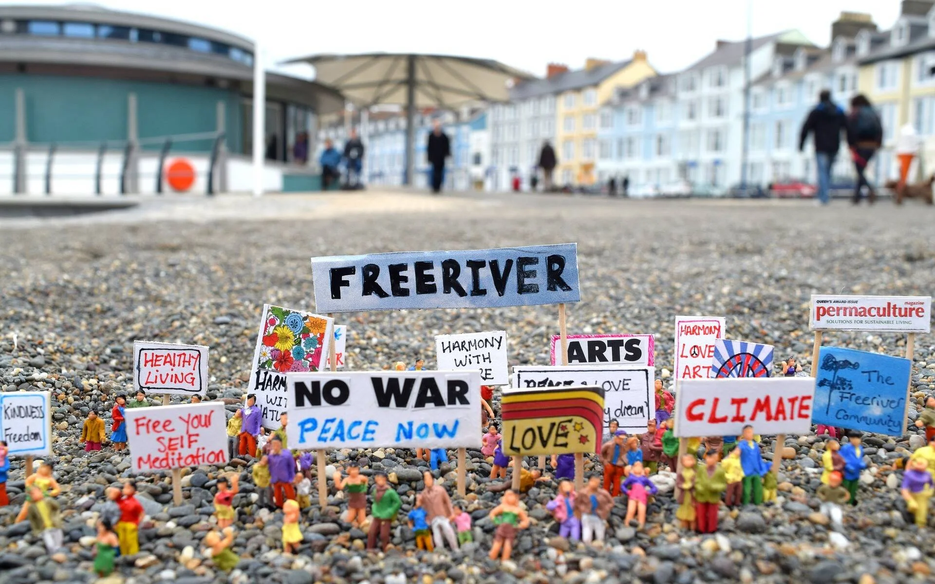 Pequeñas figuras en miniatura sobre una playa de guijarros sostienen carteles con mensajes de paz, clima y libertad, con un paseo marítimo al fondo.