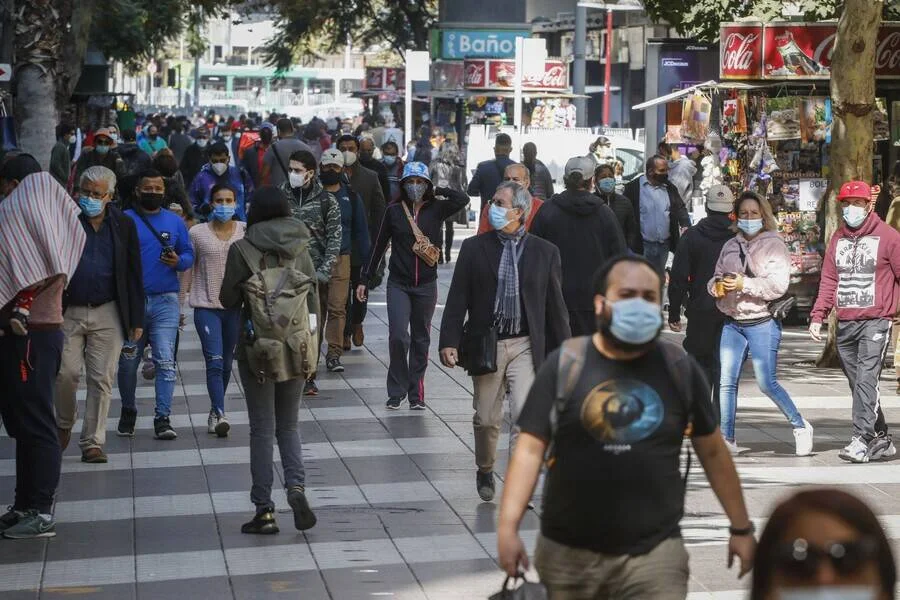 Numerosas personas con mascarillas caminan por una concurrida calle peatonal con locales comerciales al fondo.