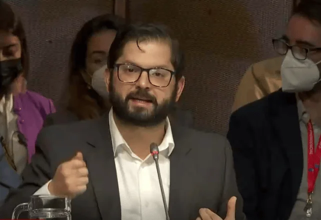 Hombre joven con barba, anteojos y saco gris habla ante un micrófono de mesa en un evento público.
