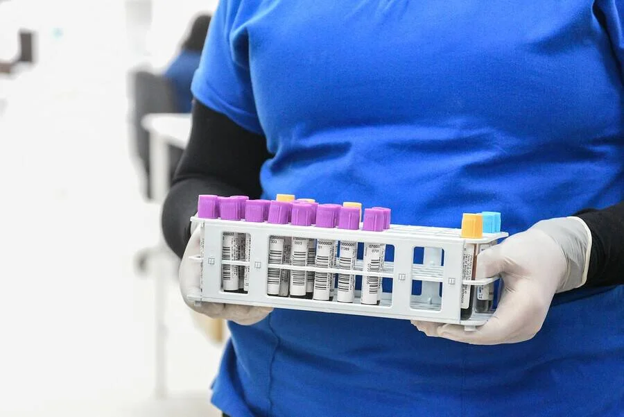 Personal sanitario con guantes sostiene una gradilla con tubos de muestras de sangre etiquetados en un laboratorio.