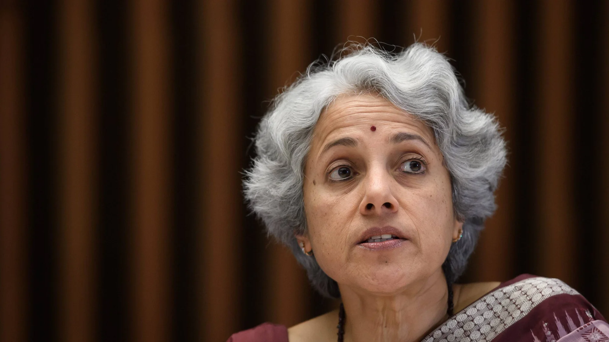 Mujer de cabello gris con bindi rojo y sari burdeos habla en un evento formal ante fondo de paneles de madera.