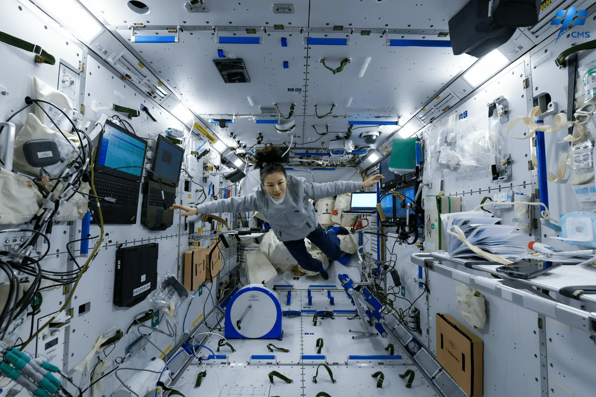 Una taikonauta flota con los brazos extendidos en el interior de la estación espacial china Tiangong, rodeada de equipos y computadoras.