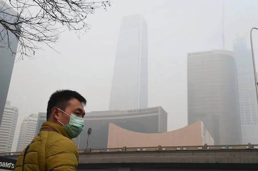 Hombre con mascarilla quirúrgica frente a rascacielos envueltos en smog en una ciudad china.