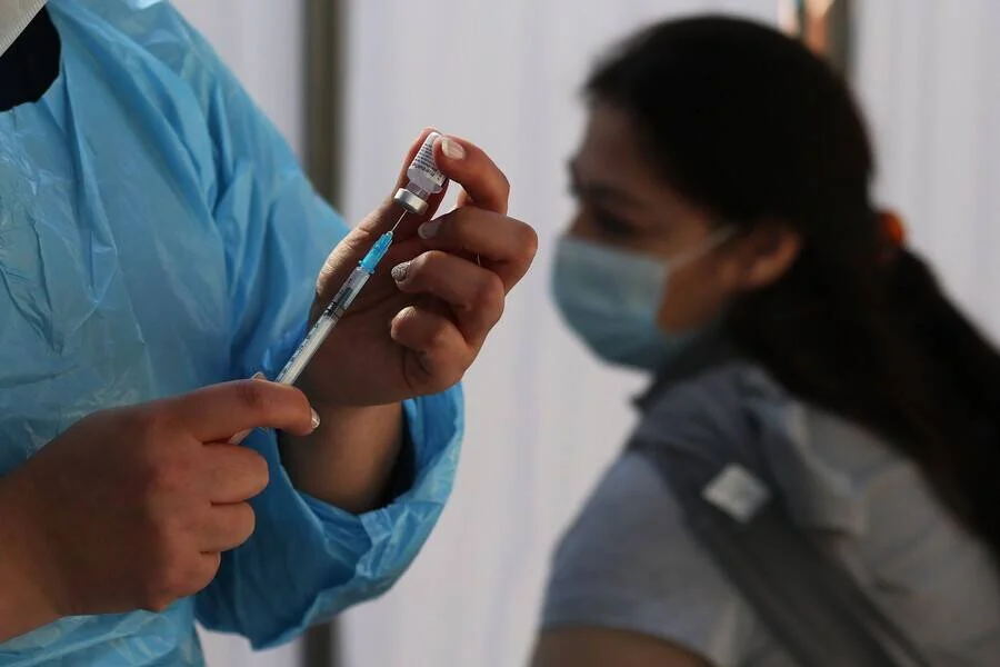 Personal sanitario con bata azul prepara una jeringa con vacuna mientras una mujer con barbijo espera para ser inoculada.
