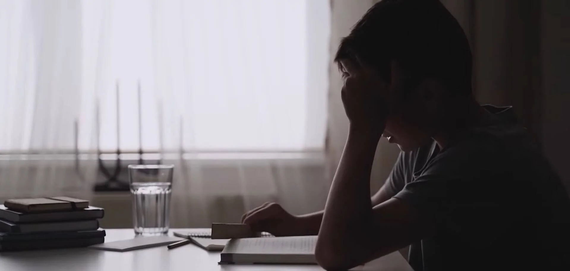 Joven con la cabeza apoyada en la mano, sentado ante una mesa con libros y un vaso de agua, en actitud de agotamiento o estrés.