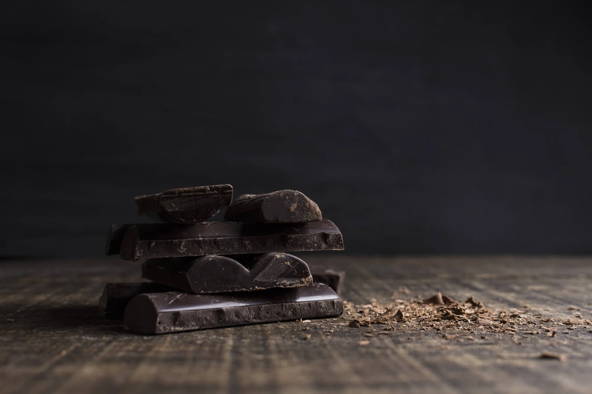 Varios trozos de chocolate negro oscuro apilados sobre una superficie de madera rústica, con virutas de cacao esparcidas alrededor.